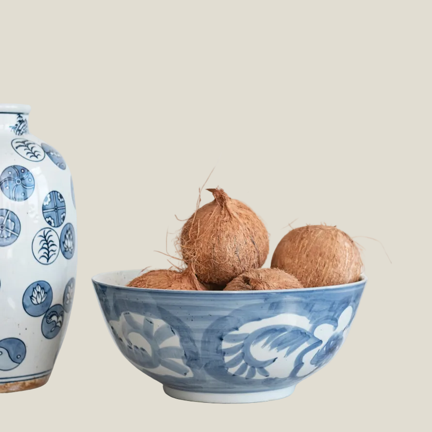 The Hampton Bloom Bowl filled with coconuts beside a blue and white vase, styled on a white tabletop
