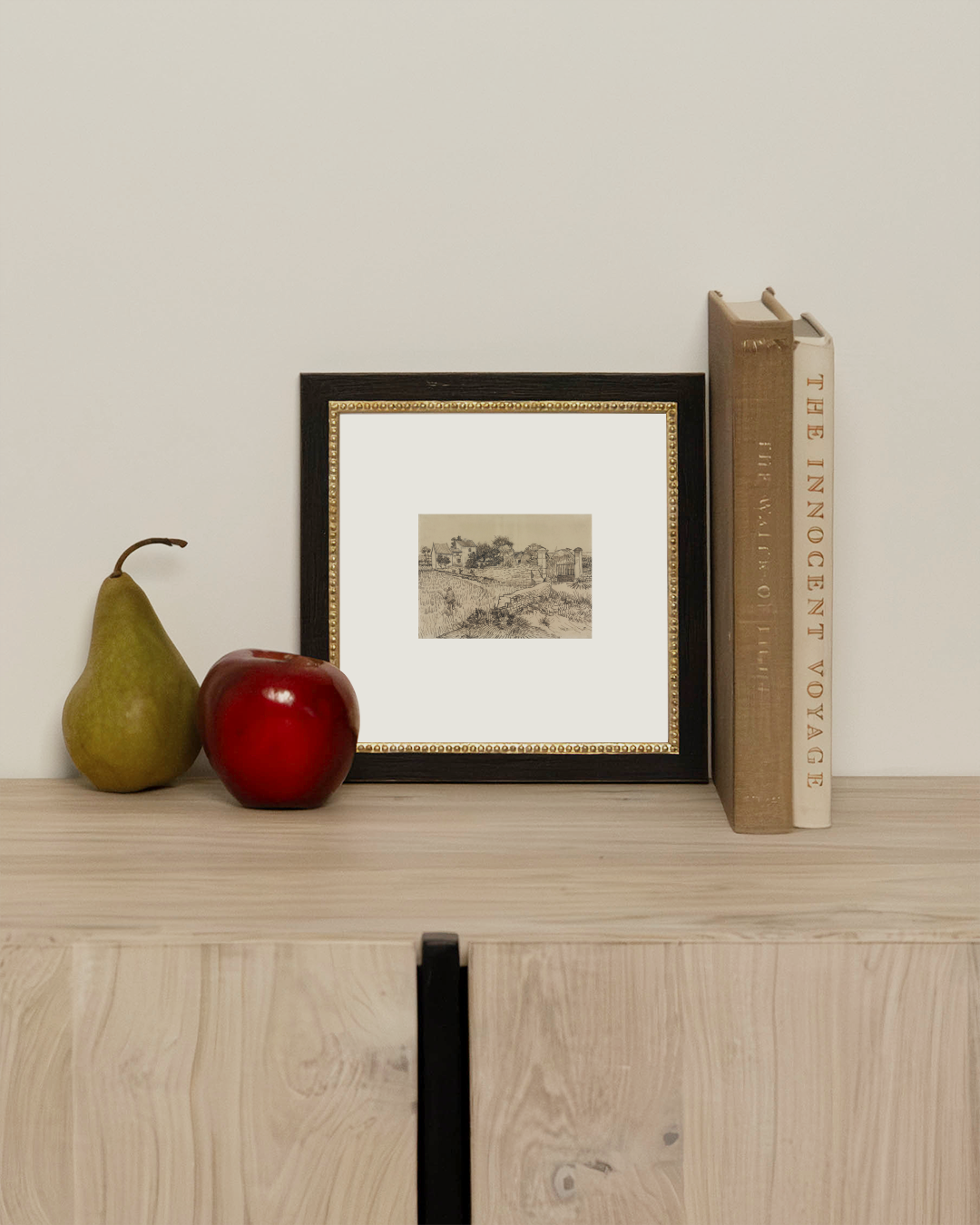 The Tuscan Village framed sketch art print leaning on a light oak cabinet beside a pear, a red apple and stacked books