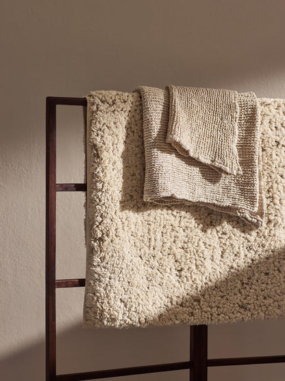 The Elsa off-white cotton and linen bath mat draped over a dark wood towel ladder in warm sunlight