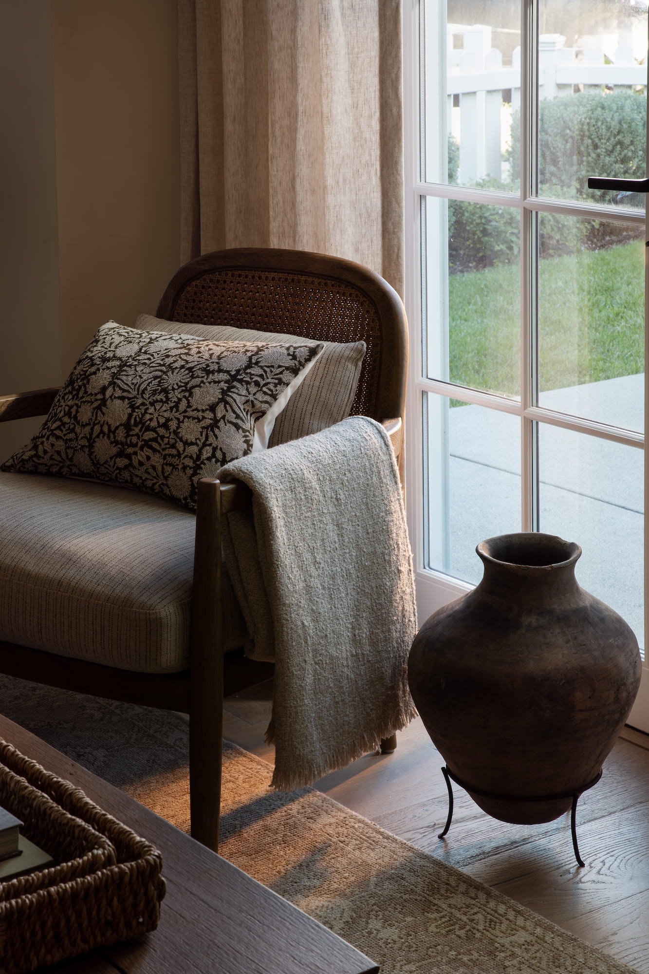 Cane-back armchair with block-print pillow and linen throw beside a glass door at dusk — Joey Snow Design Co.