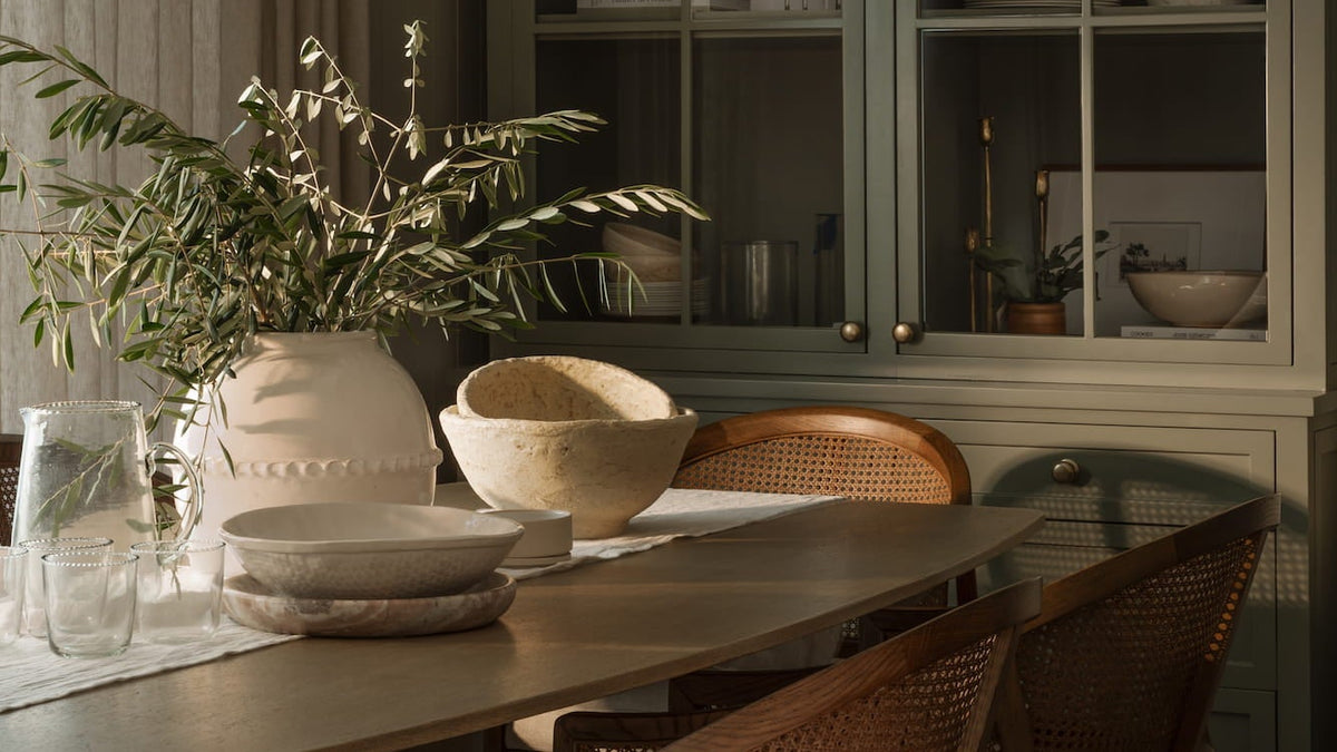 Dining table set with stone bowls and glass pitchers before a sage-green china cabinet in low evening light — Joey Snow Design Co.