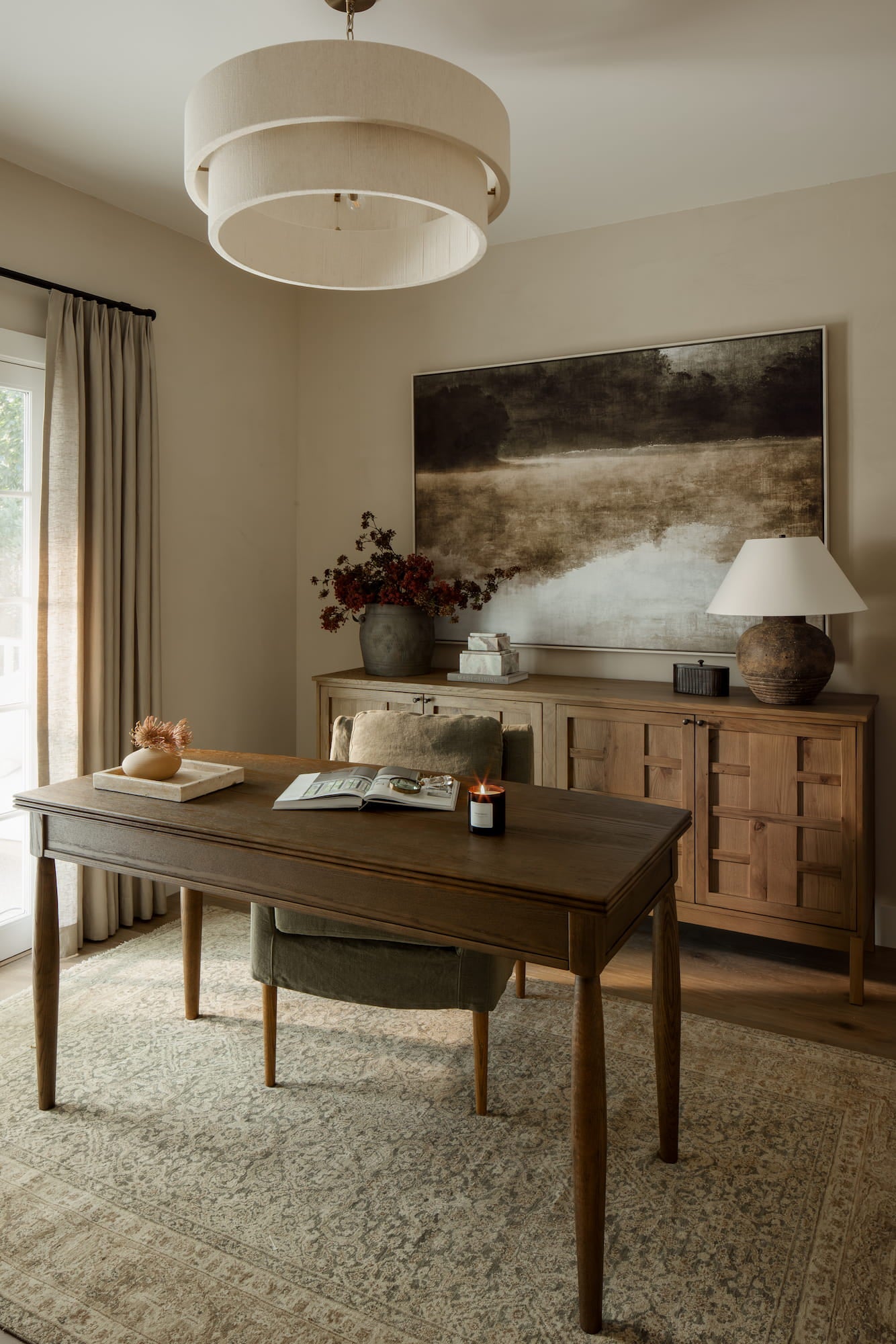 Neutral home office with wood desk, textured pendant light, large landscape artwork, and wood credenza.