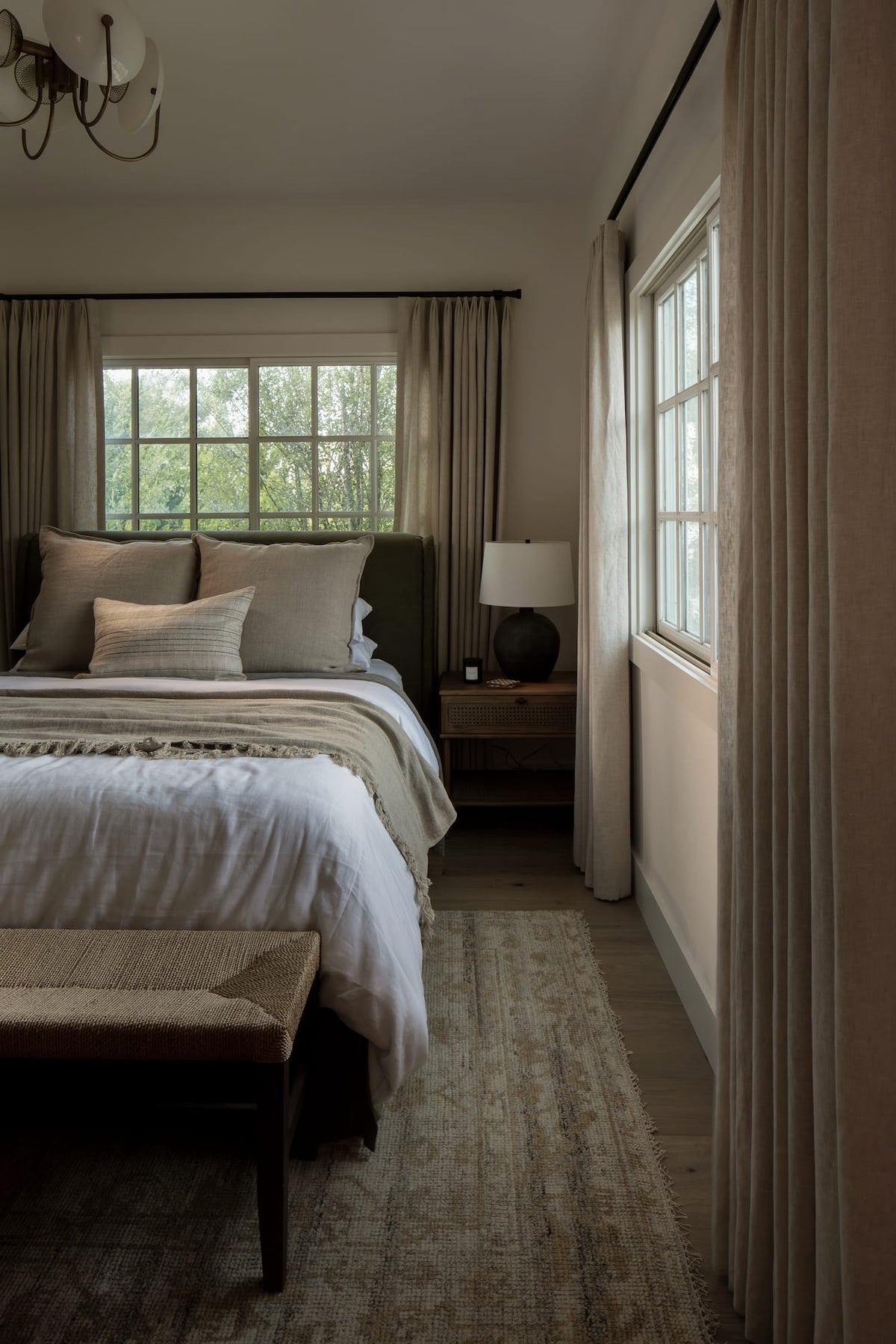 Bedroom with layered linen bedding and an olive-green headboard in soft window light — Joey Snow Design Co.