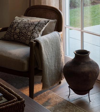 The Juliette ivory cotton bouclé throw draped over a woven armchair beside a dark ceramic vase