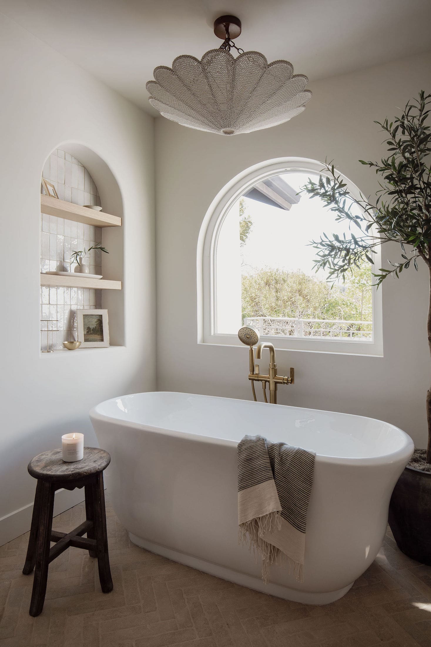 Freestanding white soaking tub beneath arched window with brass floor-mounted tub filler and woven pendant light overhead.