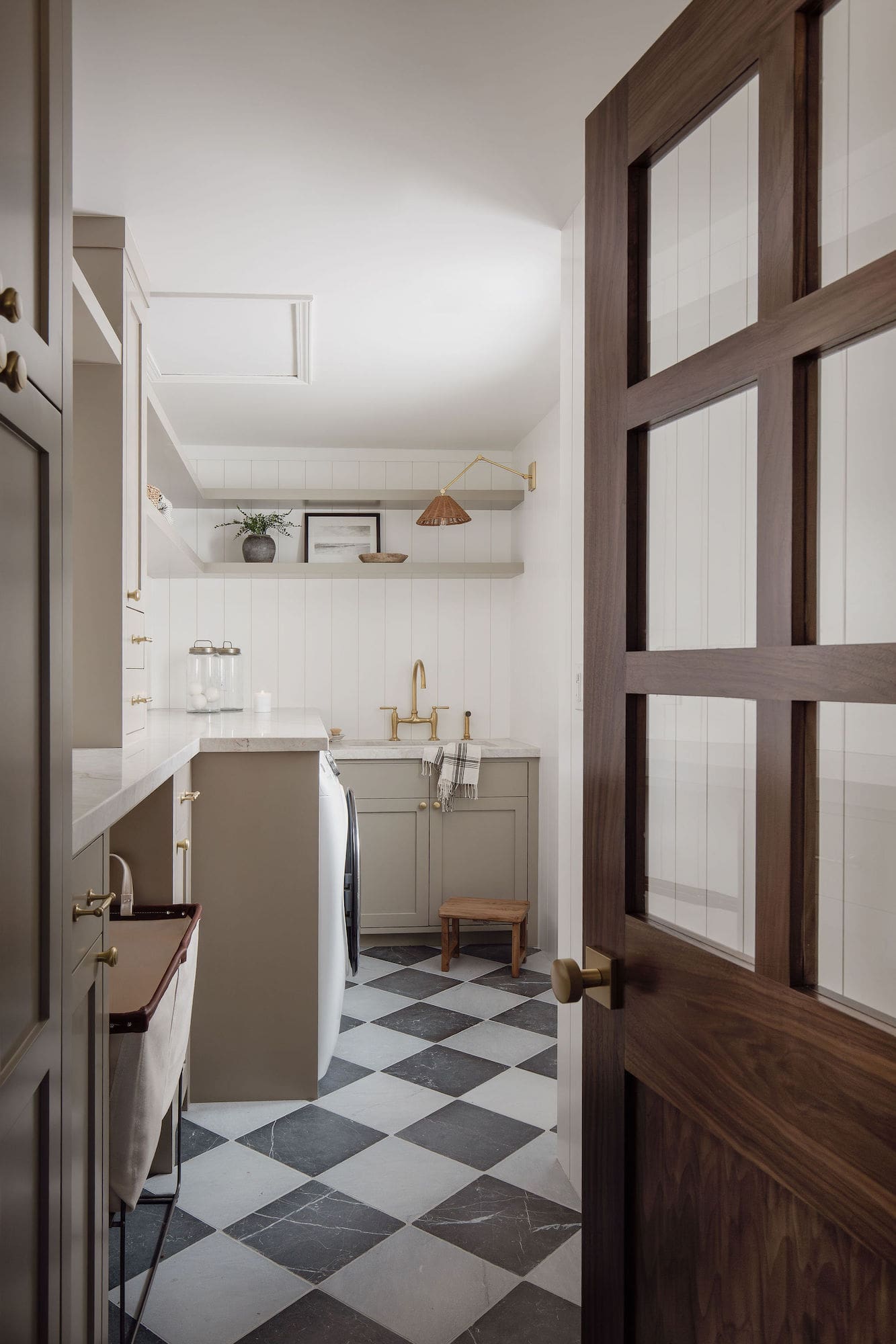 Laundry room viewed through wood-framed glass door with checkered tile floor, marble countertops, and brass hardware.